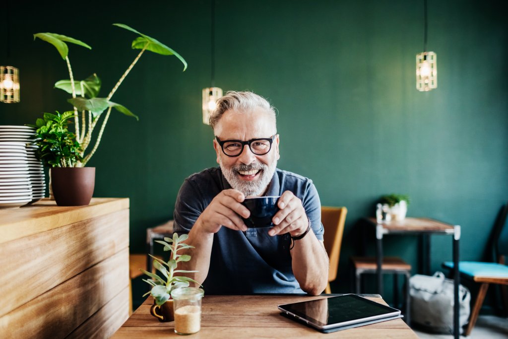 Oudere man zit aan werktafel met koffie en een tablet.